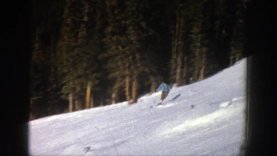 A distant skier in a blue jacket moves across the Aspen slope, with tall evergreen trees and soft 8mm exposure in the background.