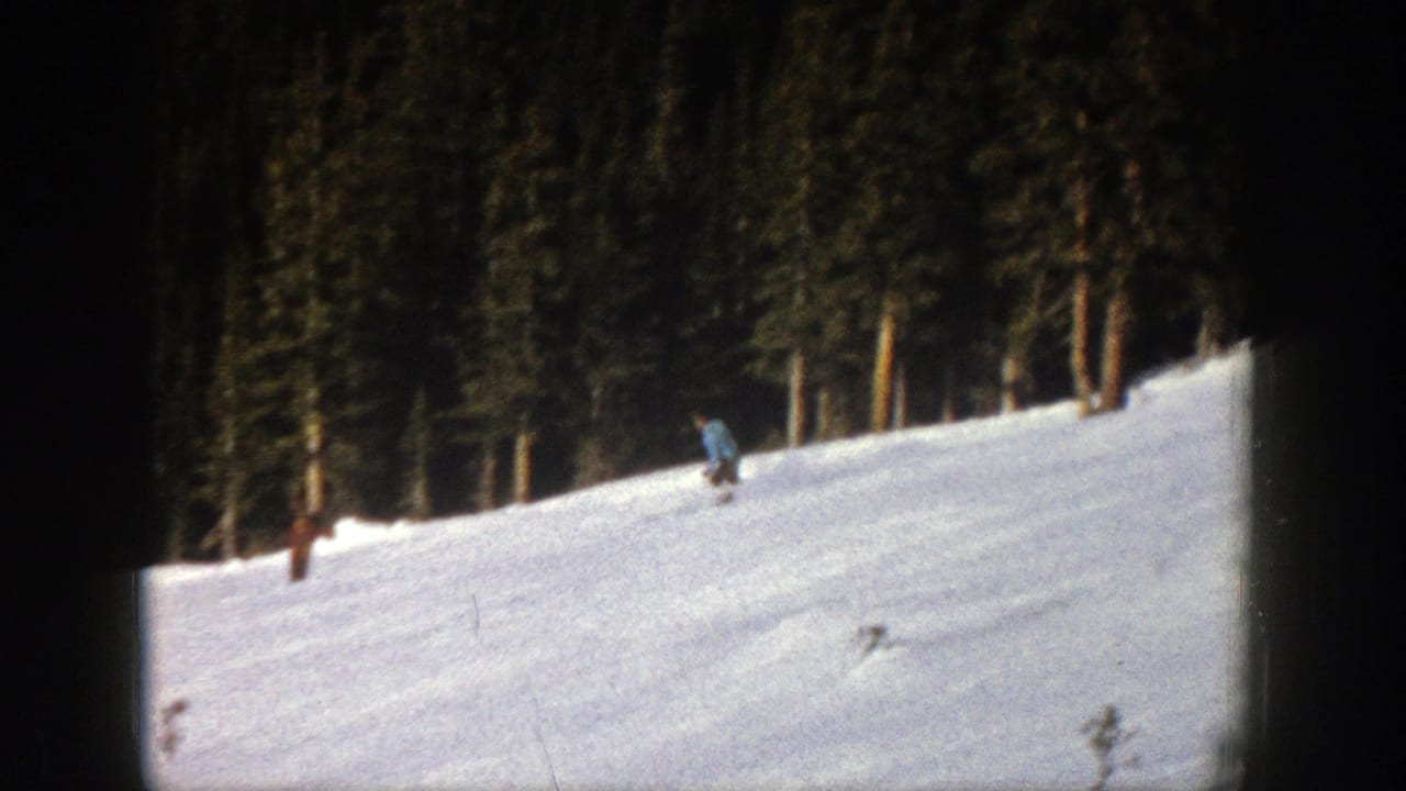 1961 Aspen home-movie frame showing a skier moving across a snow-covered slope below dense evergreen trees.