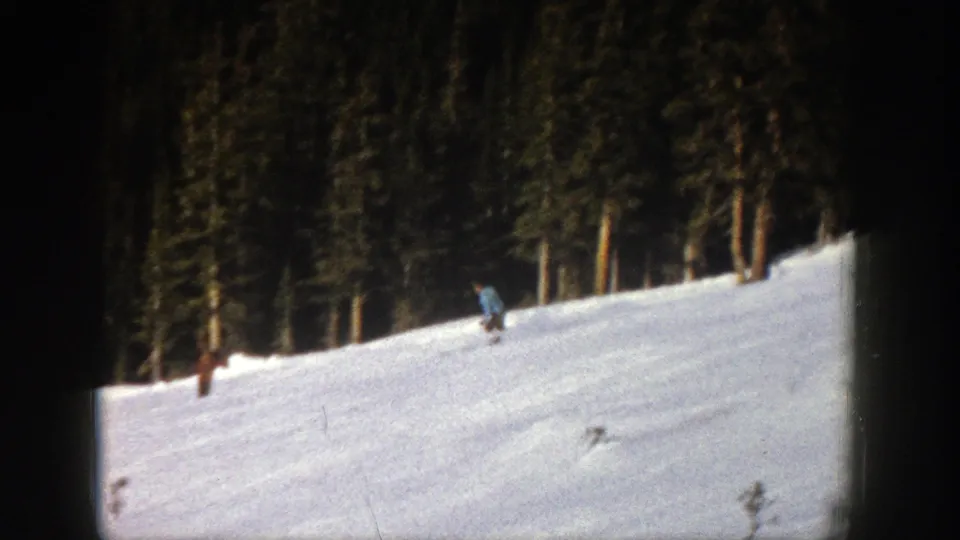 1961 Aspen home-movie frame showing a skier moving across a snow-covered slope below dense evergreen trees.