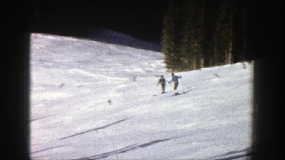 1961 Aspen home-movie frame showing several skiers traversing a broad snowy slope with evergreens at the side.