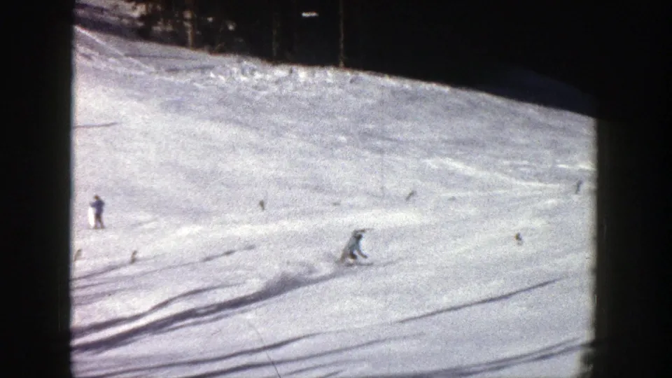 1961 Aspen home-movie frame showing several small skiers moving across a sunlit snowy mountain slope near evergreen trees.