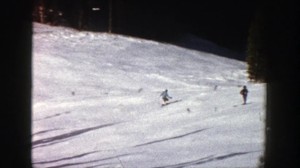 1961 Aspen footage frame with one skier crossing the snowy run and several distant figures near the tree line.
