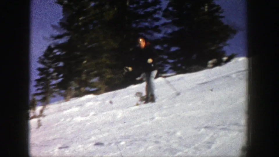1961 Aspen home-movie frame showing a skier in dark winter clothing near a snowy rise and evergreen trees.