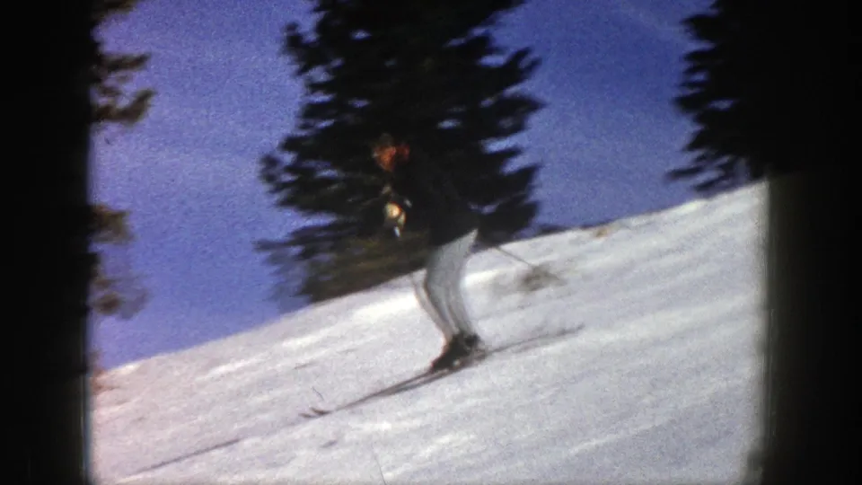 Adult skier in Aspen, Colorado, 1961 footage glides downhill on a snowy slope bordered by tall evergreens.
