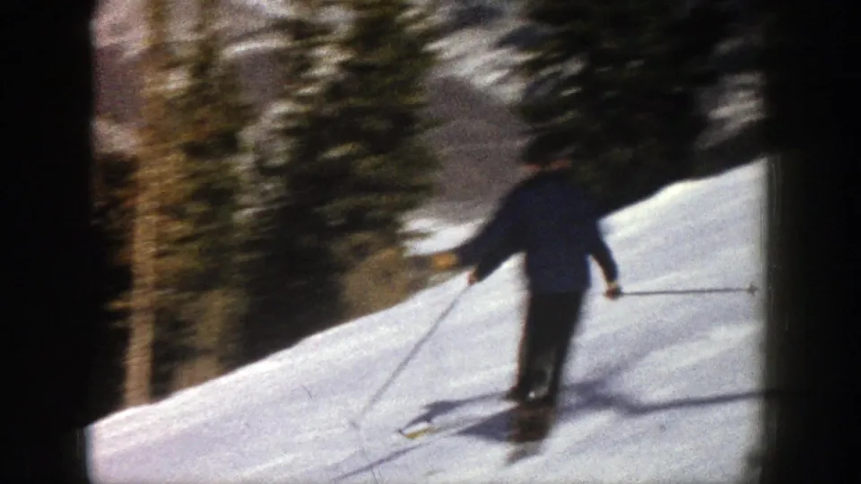 Handheld 8mm frame shows the skier from behind as he descends an Aspen slope beside evergreens.