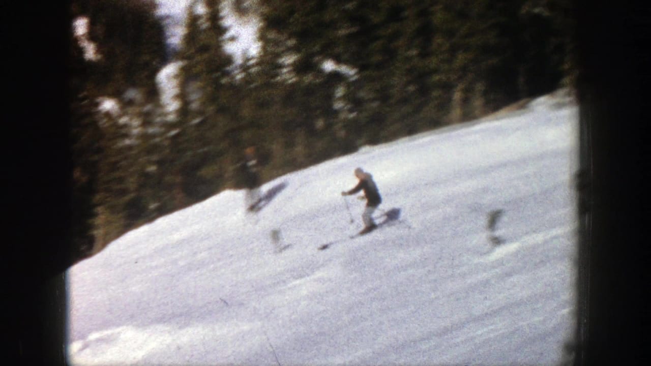 1961 Aspen home-movie frame showing a skier moving downhill on a snow-covered slope bordered by conifer forest.