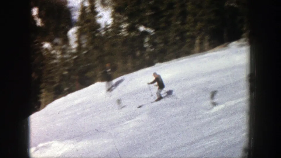 1961 Aspen home-movie frame showing a skier moving downhill on a snow-covered slope bordered by conifer forest.
