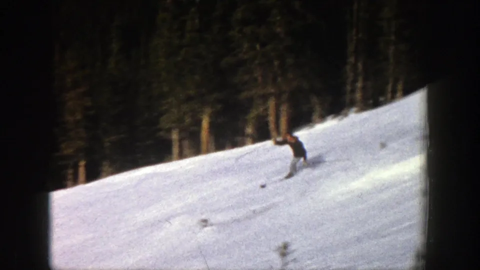 1961 Aspen frame with a skier in dark winter clothing descending across a sunlit snowy slope near a forest edge.