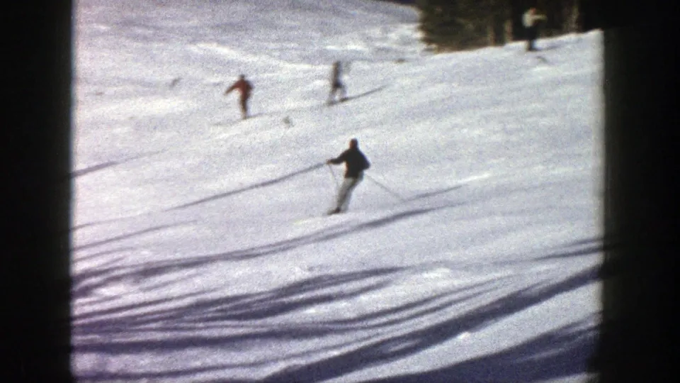 1961 Aspen frame showing multiple skiers spread across a snowy slope, with long shadows and forest along the upper edge.