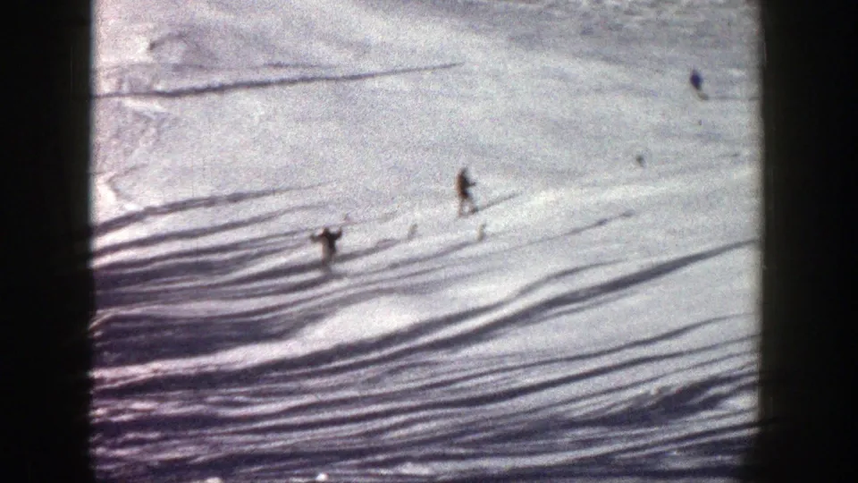 1961 Aspen home-movie frame with two visible skiers crossing a sunlit slope marked by long shadows.