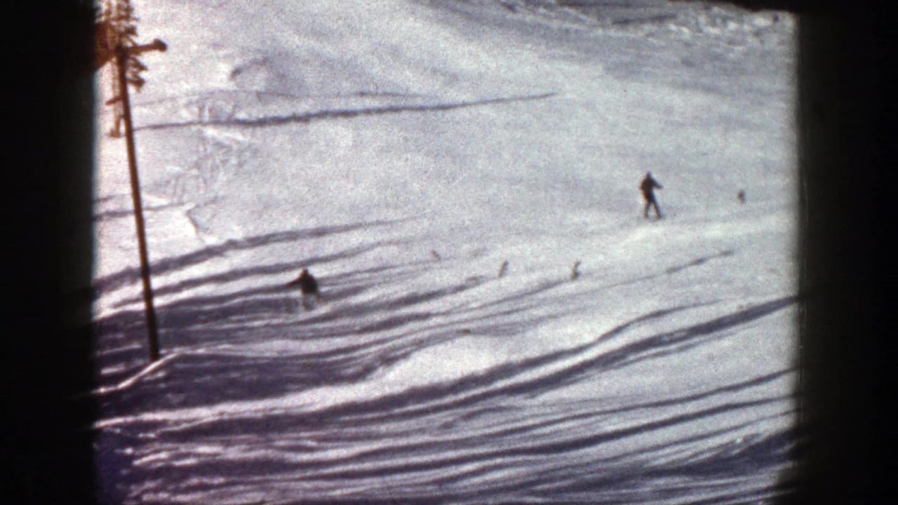 1961 Aspen 8mm frame showing a distant skier moving across a sunlit snowy slope bordered by pine trees and shadowed snow.