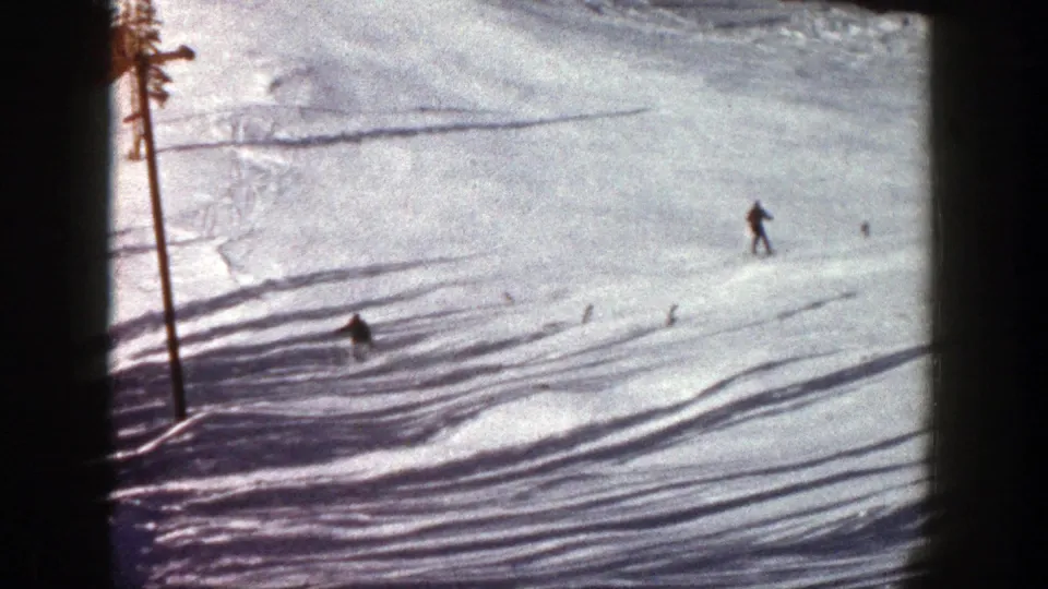 1961 Aspen 8mm frame showing a distant skier moving across a sunlit snowy slope bordered by pine trees and shadowed snow.