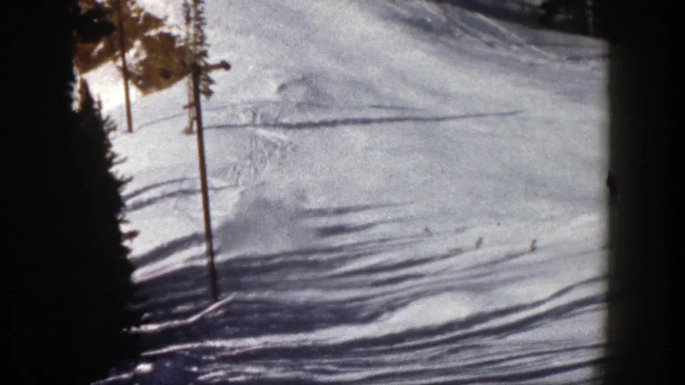 1961 Aspen ski slope seen in soft 8mm color, with a distant skier, pine trees, marker poles, and textured snow tracks.