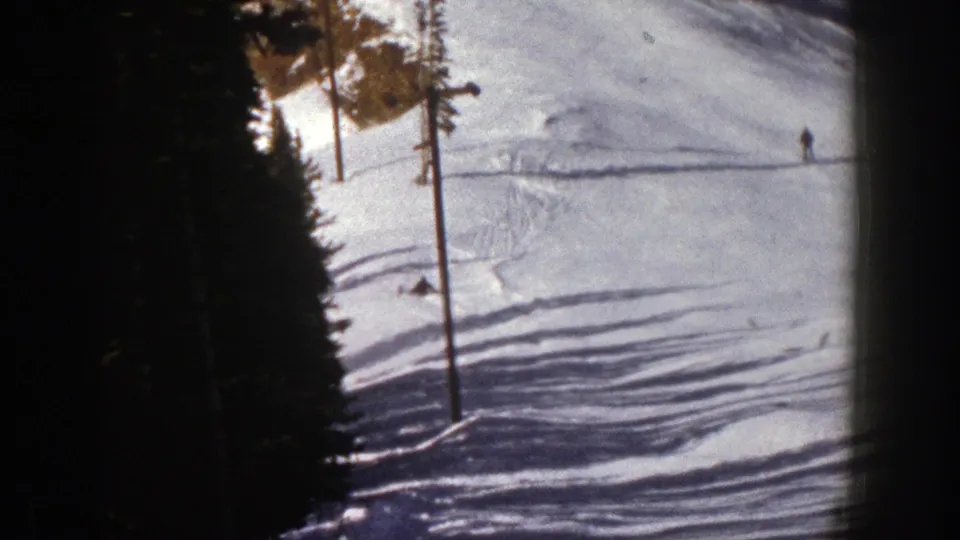 1961 Aspen home-movie frame showing a skier down on the snow below a sunlit slope, with another distant figure farther uphill.