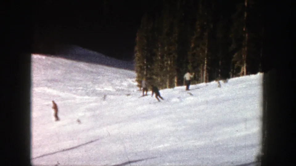 1961 Aspen home-movie frame showing a skier near evergreens and another distant skier on a bright, softly focused slope.