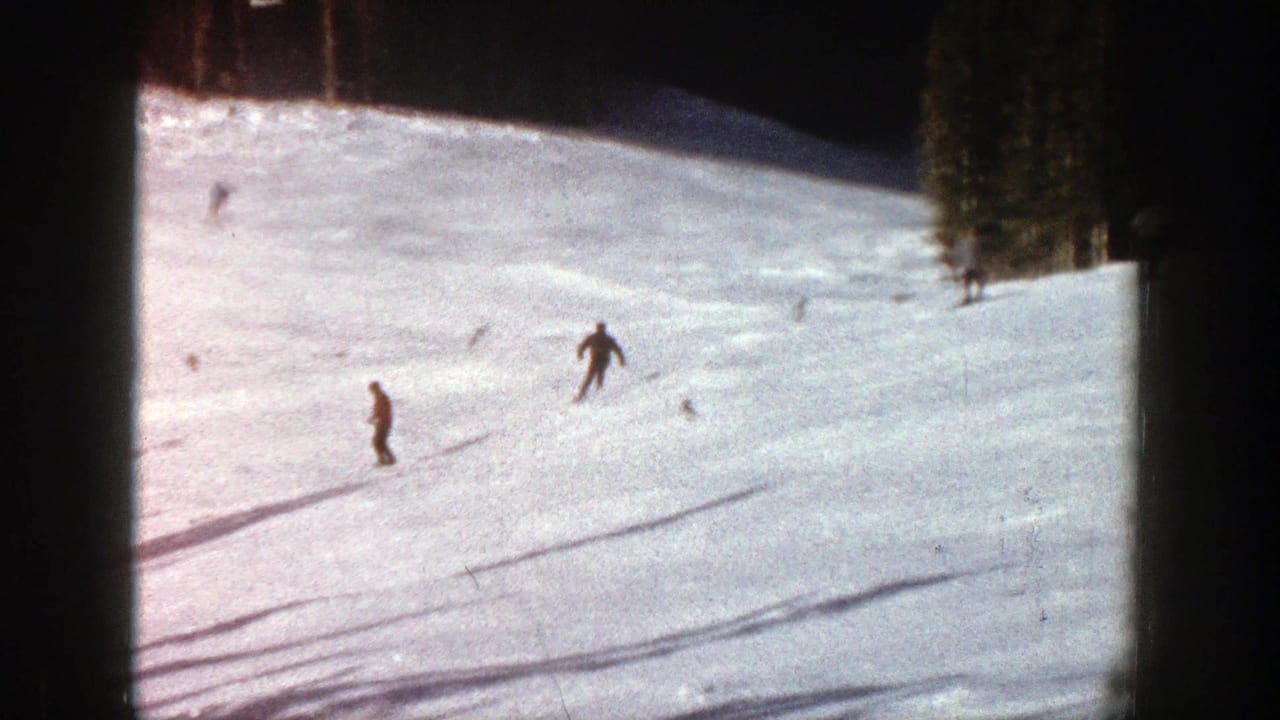 1961 Aspen home-movie frame showing distant skiers moving across a snowy slope with evergreen trees at the side.