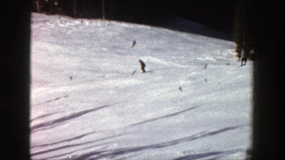 1961 Aspen home-movie frame with a central skier descending past faint tracks and distant figures on the upper slope.