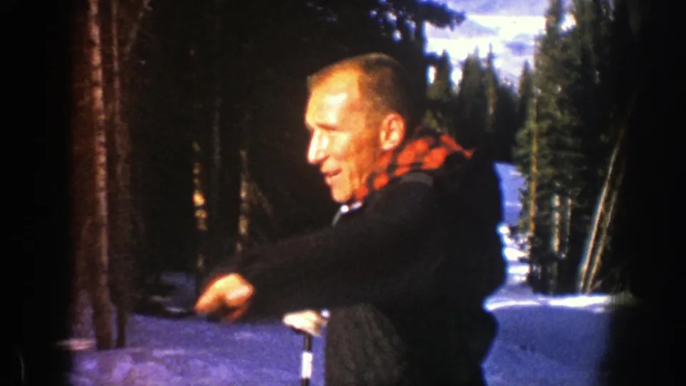 Man in a winter coat gestures near a ski pole in snowy Aspen woods in this 1961 home-movie frame.