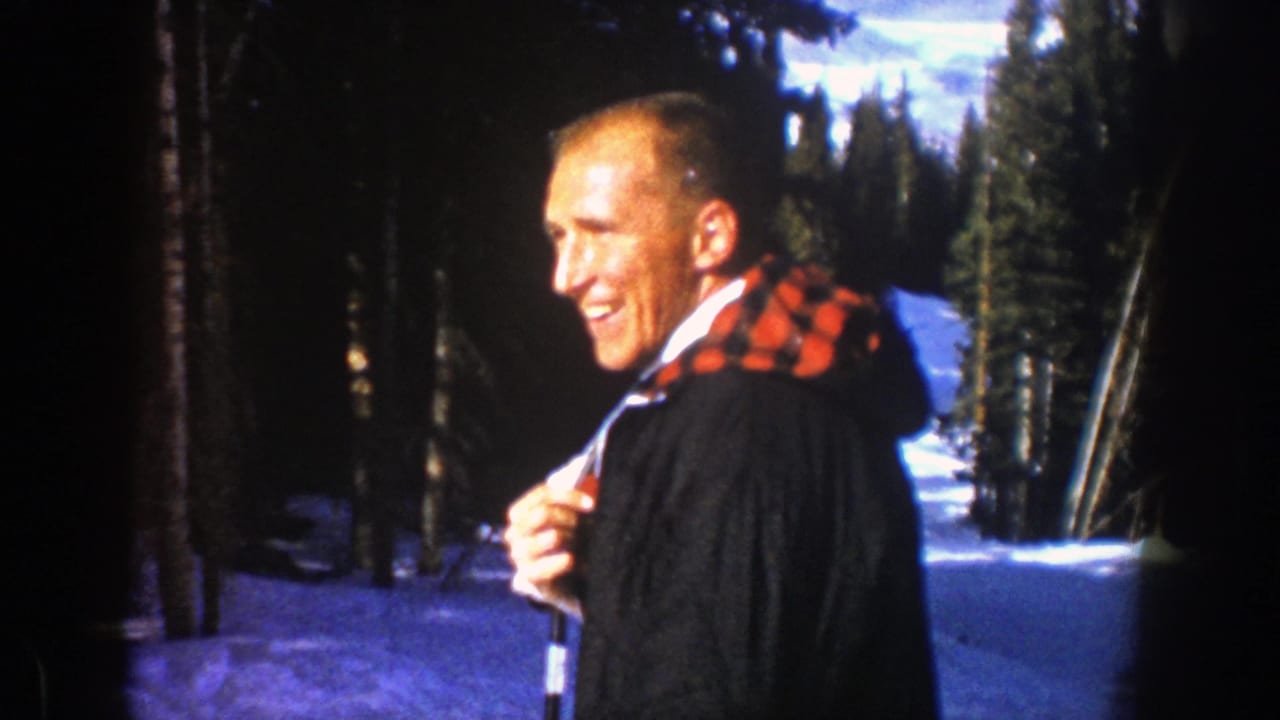 In 1961 Aspen footage, an adult man in a dark coat and red plaid scarf smiles while standing on a snowy ski slope.