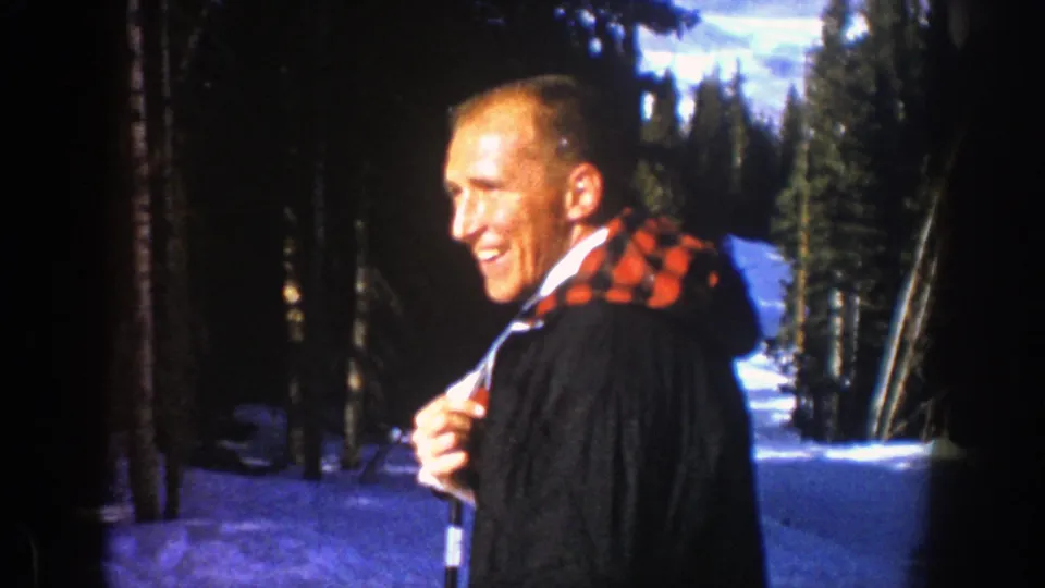 In 1961 Aspen footage, an adult man in a dark coat and red plaid scarf smiles while standing on a snowy ski slope.