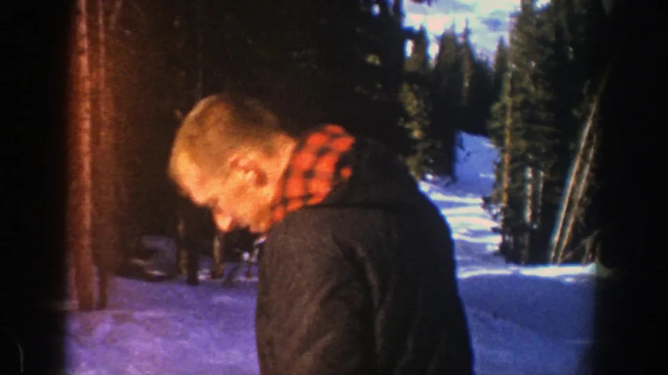 An adult skier in Aspen appears in profile, wearing a dark coat and red plaid scarf beside snow and tall evergreens.