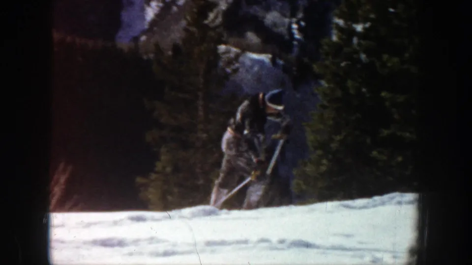 A bundled figure, possibly on snowshoes or skis, is seen near trees and rock on the snowy Aspen terrain.