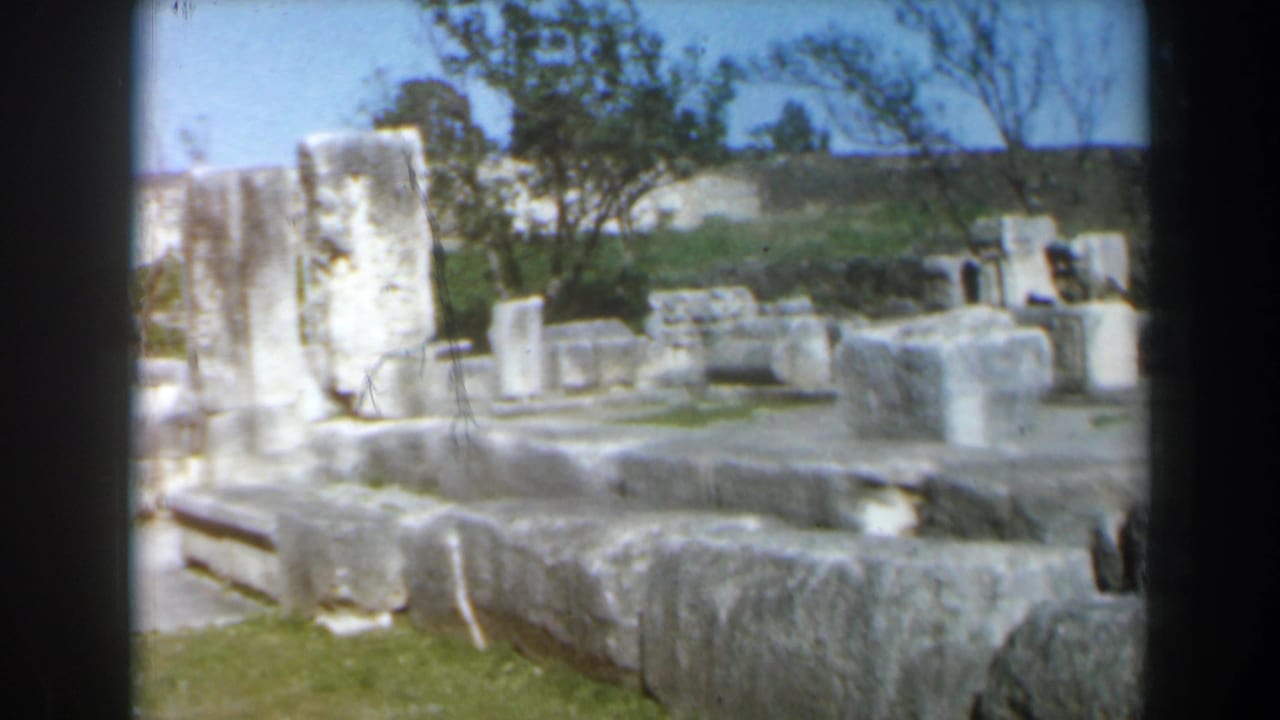 1960 Israel 8mm frame showing weathered stone ruins, low walls, upright fragments, grass, trees, and bright sky at an outdoor site.
