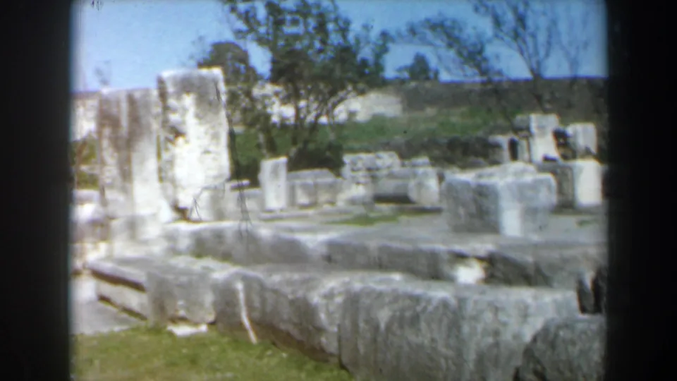1960 Israel 8mm frame showing weathered stone ruins, low walls, upright fragments, grass, trees, and bright sky at an outdoor site.