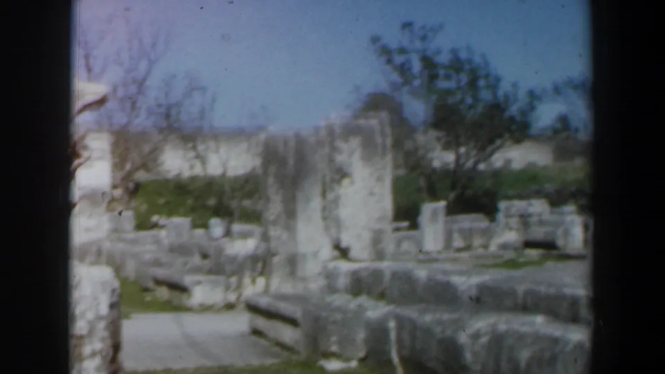 1960 Israel 8mm view of an archaeological ruin area with upright stone fragments, low walls, grass, trees, and open sky.