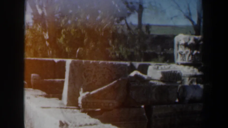 1960 Israel 8mm frame with carved stone blocks, broken architectural fragments, a paved path, trees, and sun flare.