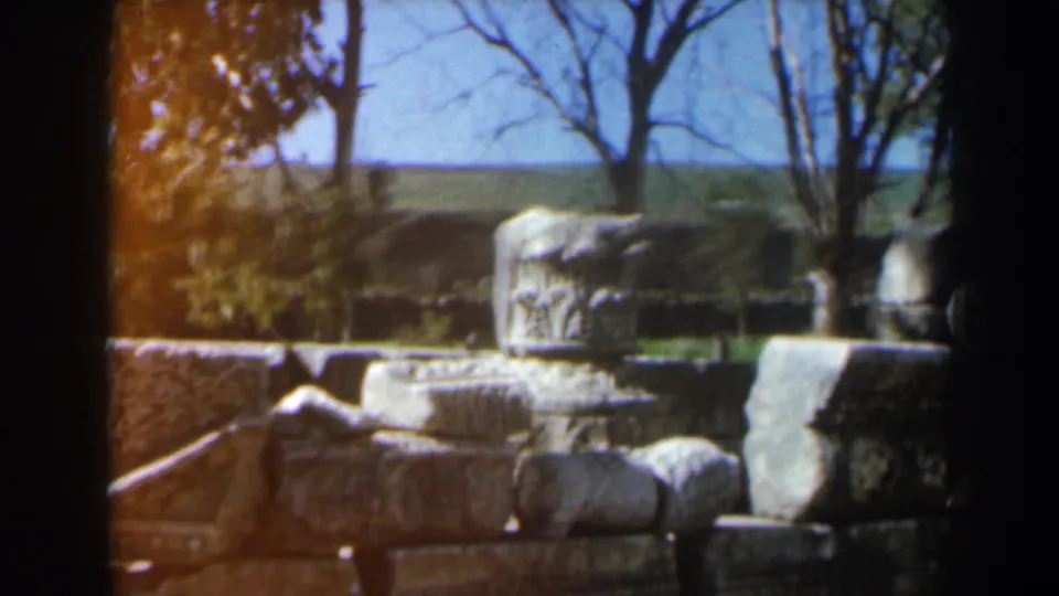 1960 Israel 8mm view of a carved stone block among scattered ruins, with trees, grass, and open ground behind it.
