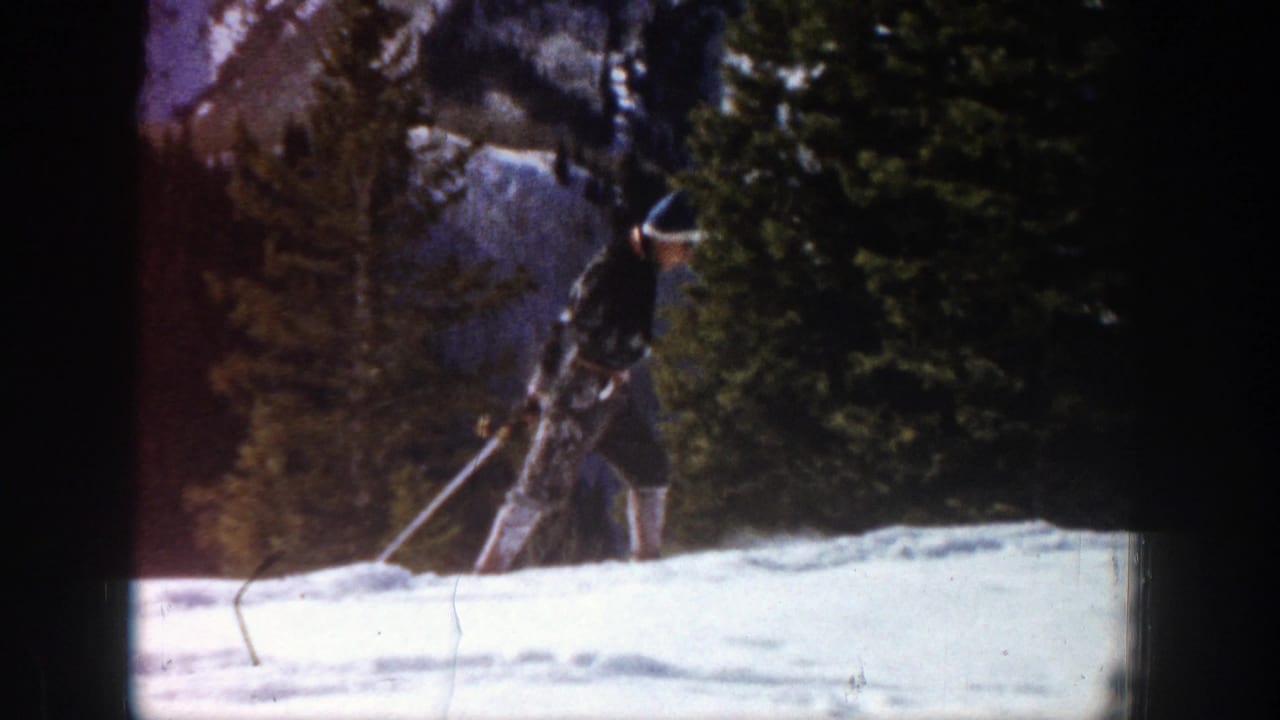 In Aspen, Colorado in 1961, a skier in a black hat and dark jacket moves uphill across snow beside evergreen trees.