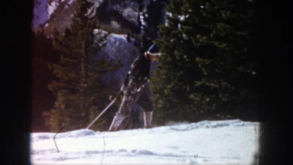 In Aspen, Colorado in 1961, a skier in a black hat and dark jacket moves uphill across snow beside evergreen trees.