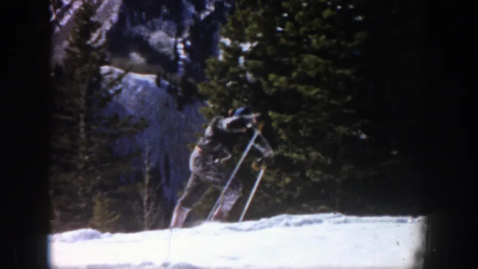 The 1961 Aspen home-movie frame shows the skier bent forward, using poles against bright snow with trees behind him.