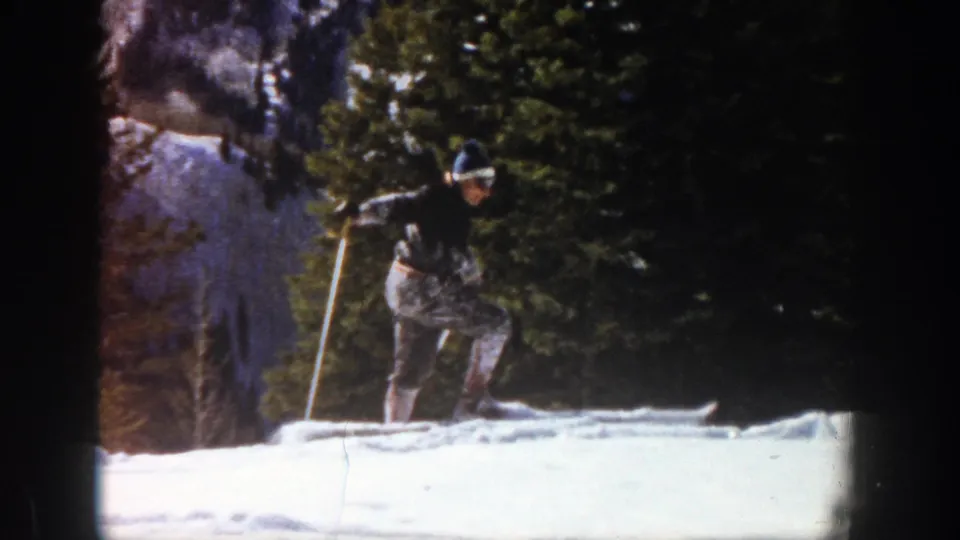 In a 1961 Aspen winter scene, the skier plants poles and lifts one ski while moving uphill through the snowy forest edge.