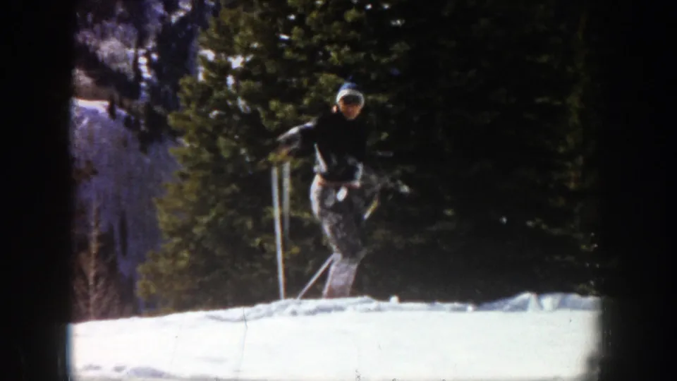 The skier appears to face the camera briefly in the 1961 Aspen footage, standing on skis with poles against a snowy wooded background.
