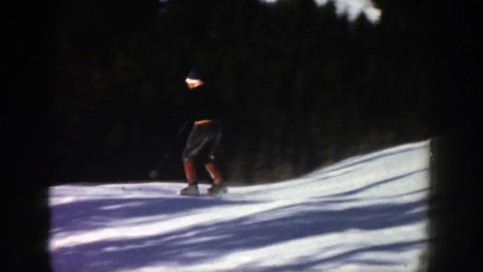 The skier crosses a shaded section of the Aspen snow slope, with strong contrast from the trees and sunlit snow.