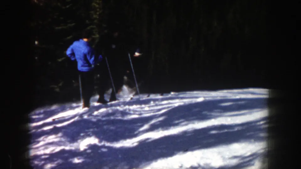 A person in a blue jacket stands on skis near another skier on the Aspen slope, with evergreen trees and bright snow visible.
