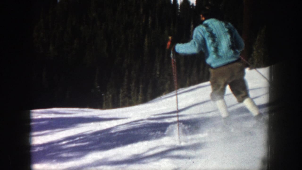 Skier in a blue sweater descends a snowy slope with evergreen trees behind.