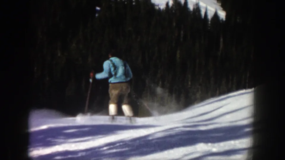 The skier moves farther down the Aspen slope, seen from behind as powder trails near the skis and evergreens fill the background.