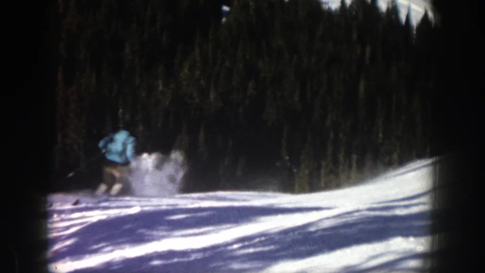 The skier is farther away near the left side of the frame, with disturbed snow and dark evergreen trees behind the white slope.