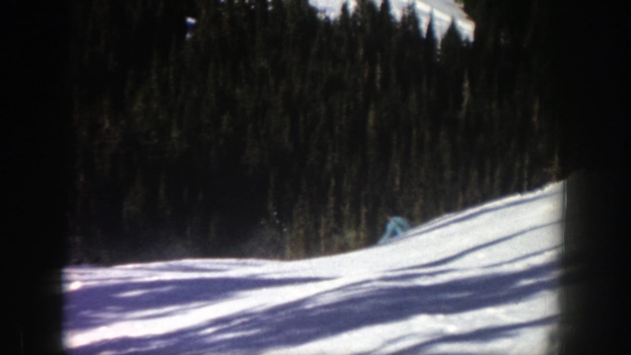 Distant skier in blue on a snowy slope bordered by dark evergreen trees.