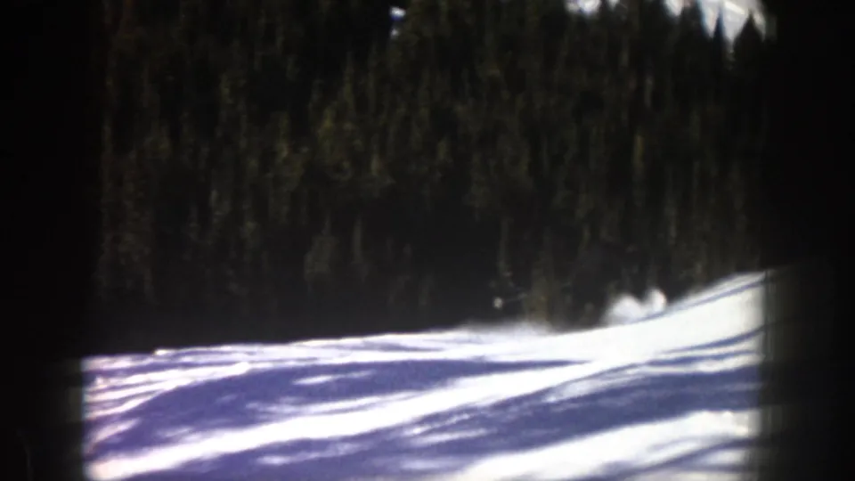 1961 Aspen ski footage frame with a snow-covered rise, forest backdrop, and a blurred trace of movement near the slope crest.
