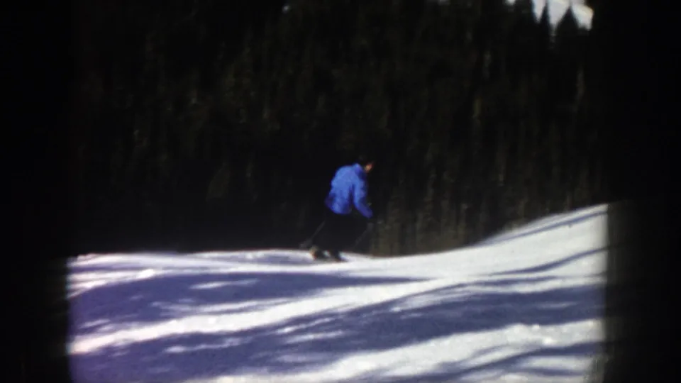 The skier continues downhill on the Aspen snow, framed against dense evergreens and strong sunlight.