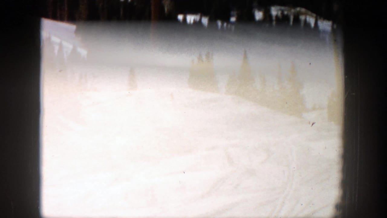Overexposed snowy ski slope with dark film borders and faint trees near the horizon.