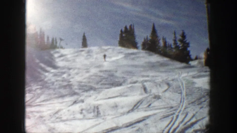 1961 Aspen frame showing a lone distant skier on a sunlit snowy hill with evergreen trees, ski tracks, and blue sky.