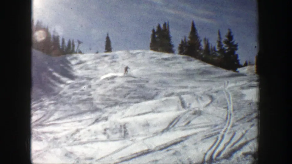 1961 Aspen home-movie frame of the skier moving across a tracked snowy hill beneath dark evergreen trees and winter sun.