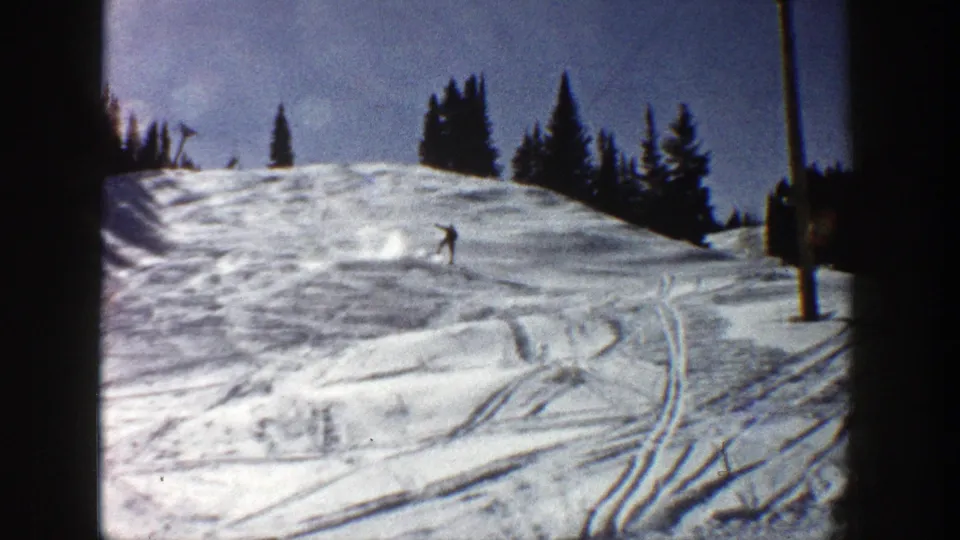 1961 Aspen frame with a distant skier descending a sunlit snowy run, evergreen trees on the ridge, ski tracks, and a pole at frame right.