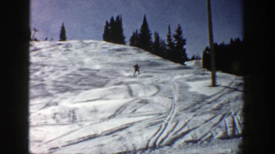 1961 Aspen home-movie frame showing a distant skier on a bright snowy slope with tree line, tracks, and dark film borders.