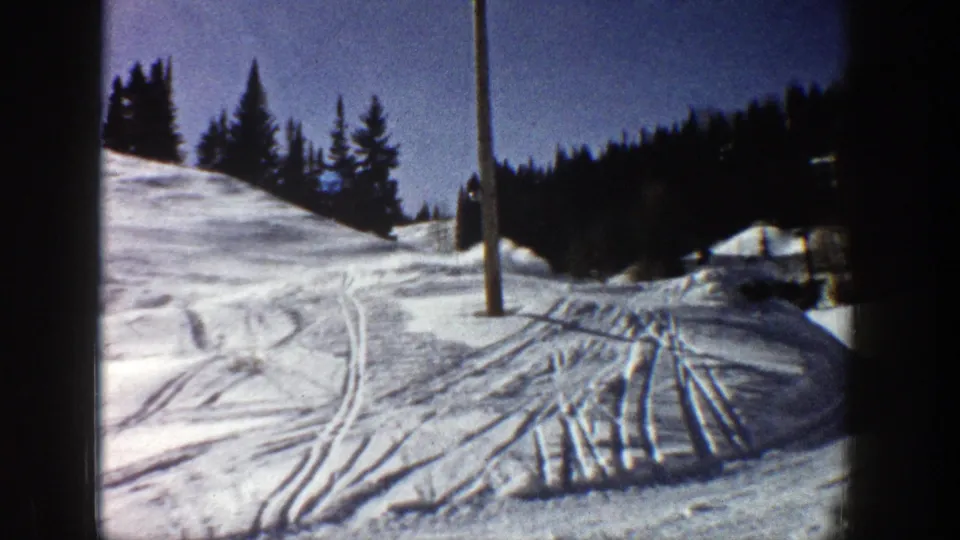 Bright 1961 Aspen ski-slope view with crisscrossing tracks, evergreen ridge, blue sky, and a pole in the snow.